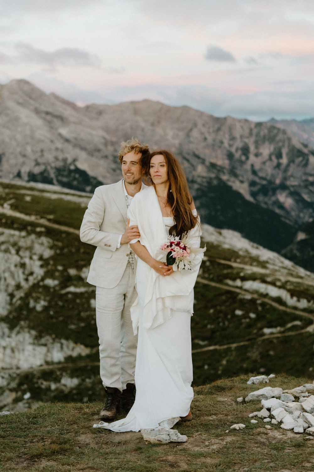 A couple in wedding attire wrap into each other and look out at the sunrise with the mountains behind them during their Italian Dolomites elopement.