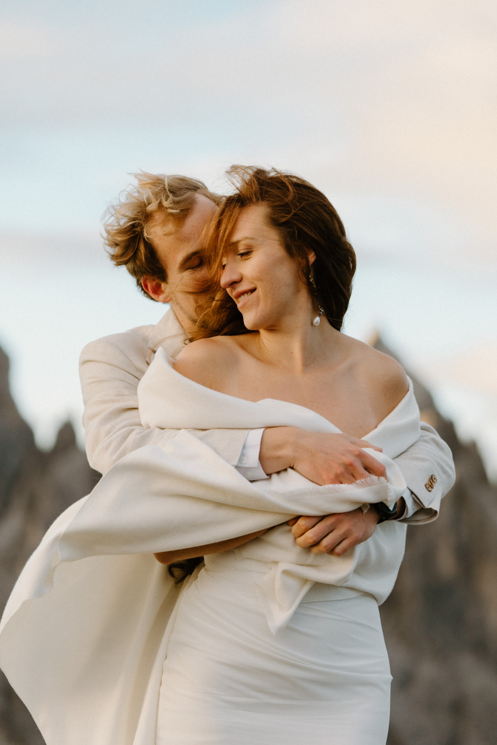 A couple in wedding attire wraps into a white shawl as the wind picks it up. The sun rises over the mountains behind them during their Italian Dolomites elopement.