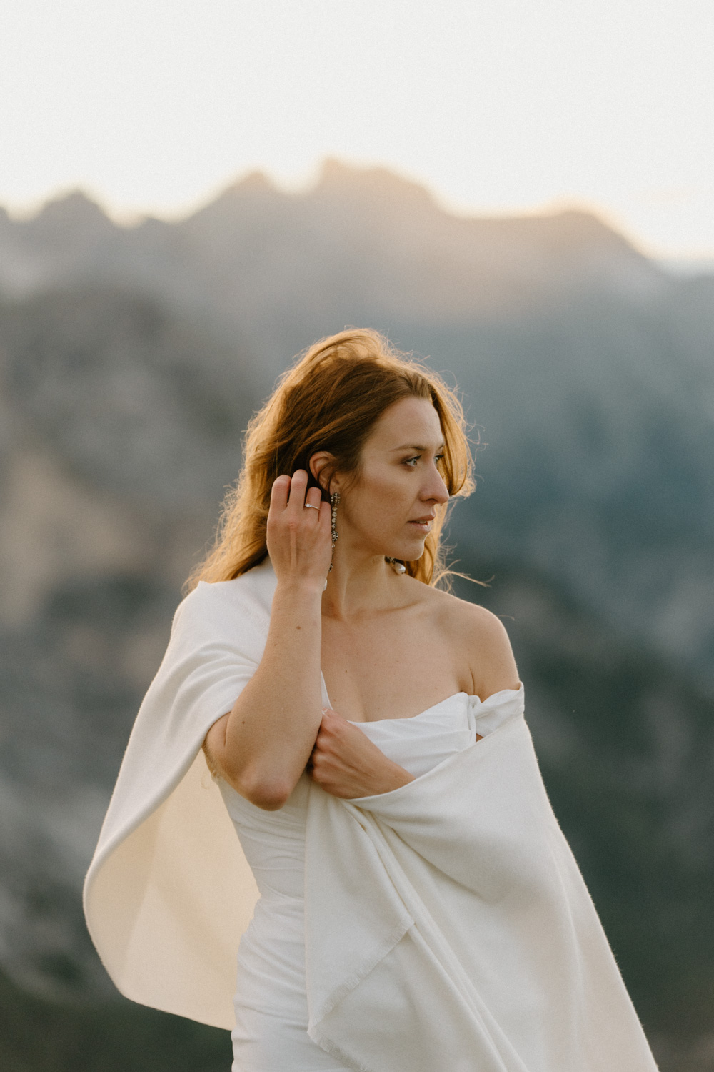 A bride wraps a white shawl around her and looks behind her at the sunrise over the mountains during her Italian Dolomites elopement.