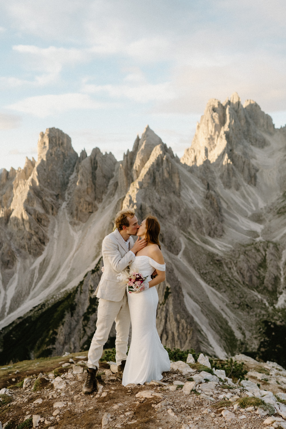 A couple in wedding attire stand together and kiss in front of Cadini di Misurina during their Italian Dolomites elopement.