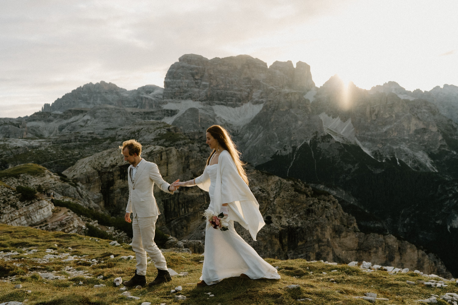 A couple in wedding attire walk hand in hand as the sun rises over the mountains behind them during their Italian Dolomites elopement.