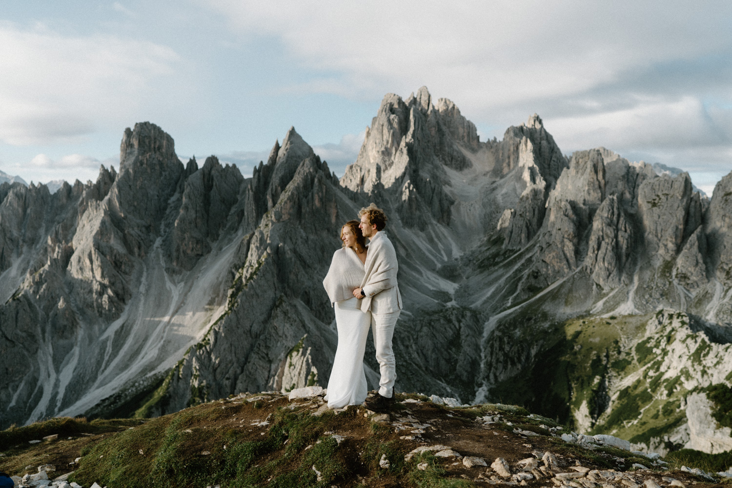 A couple in wedding attire embrace and watch the sun rise with Cadini di Misurina behind them during their Italian Dolomites elopement. 