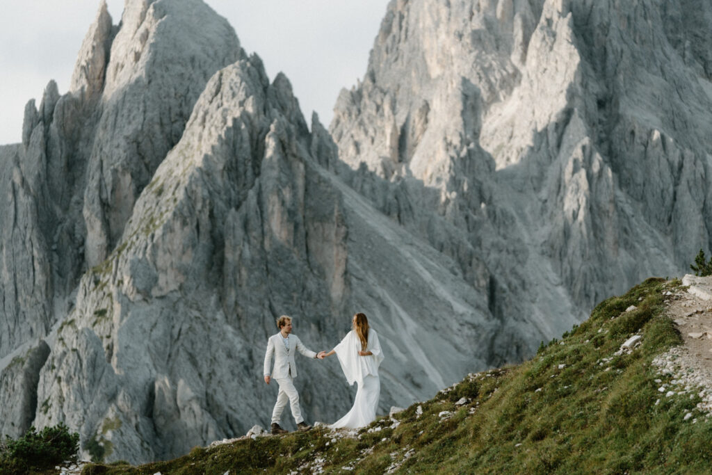 A far away shot of a couple in wedding attire, walking hand in hand in front of a gray, rugged mountain range during their Italian Dolomites elopement.