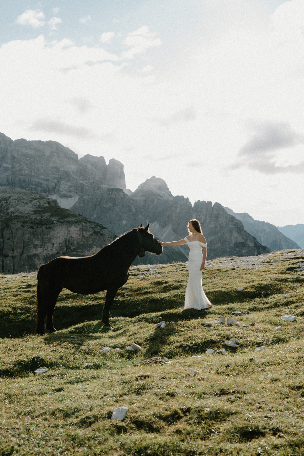 A bride walks up to a black horse and touches his nose, with the mountains behind then during her Italian Dolomites elopement.