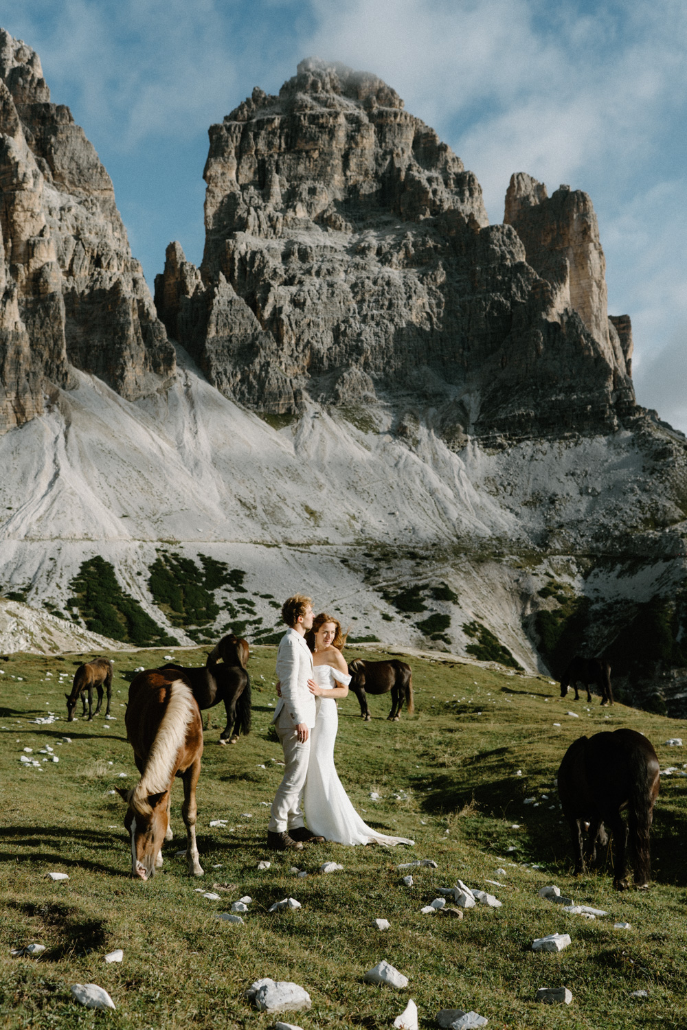 A couple in wedding attire stand in front of a mountain, surrounded by horses, during their Italian Dolomites elopement.