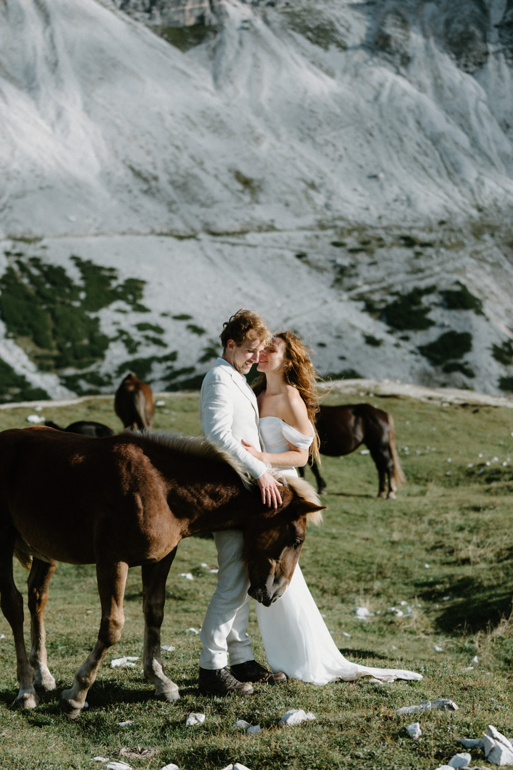 A couple in wedding attire stand in front of a mountain, surrounded by horses, during their Italian Dolomites elopement.