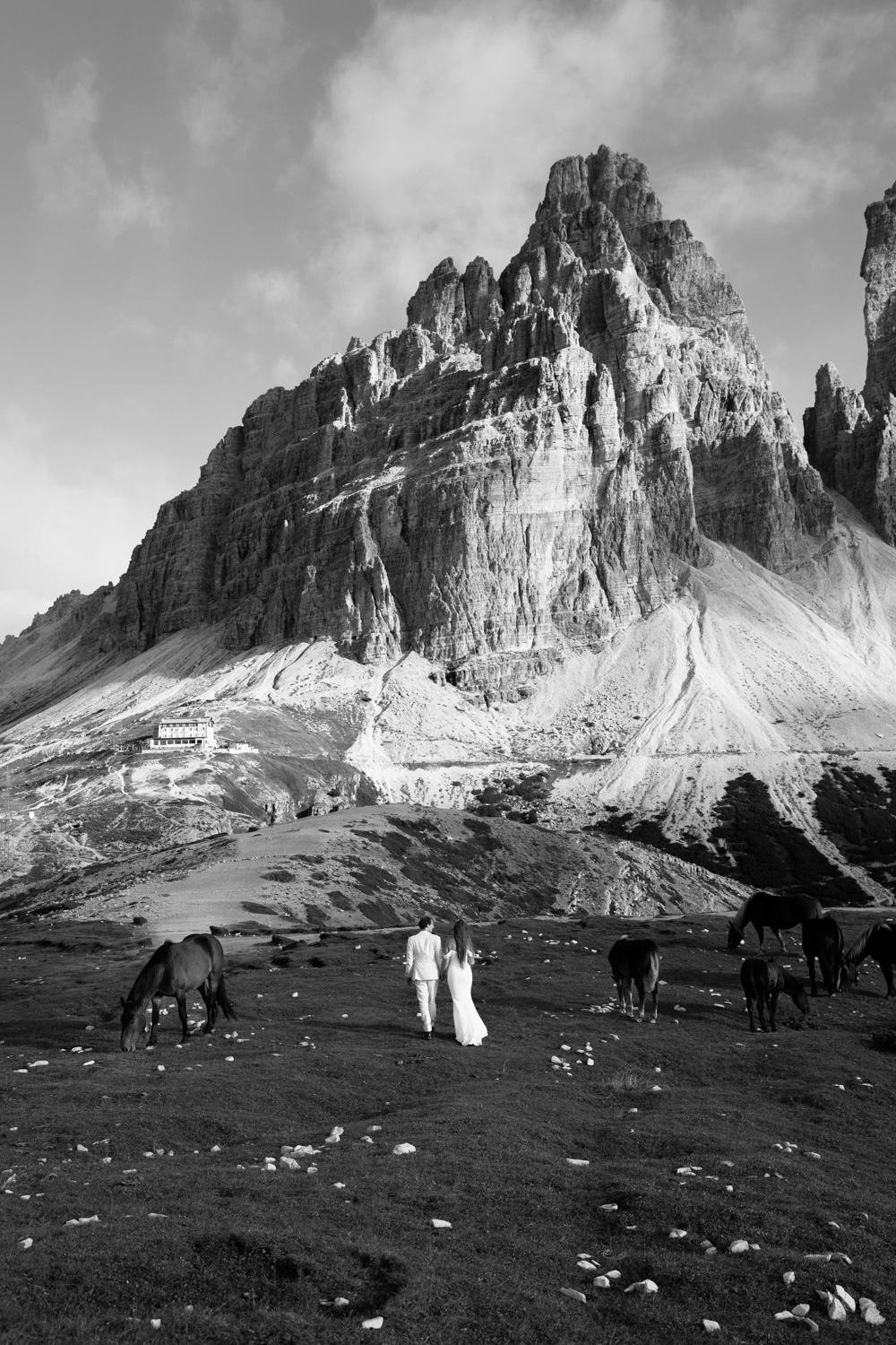 A back and white photo of a couple in wedding attire standing in front of a mountain, far away, surrounded by horses, during their Italian Dolomites elopement.