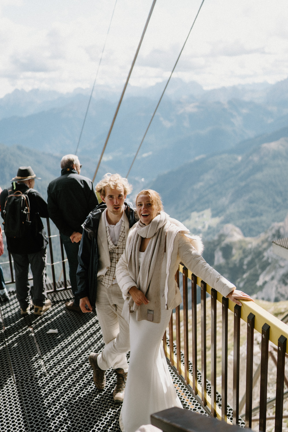 A photo of a couple in wedding attire and jackets standing and smiling at the top of a tram during their Italian Dolomites elopement. 