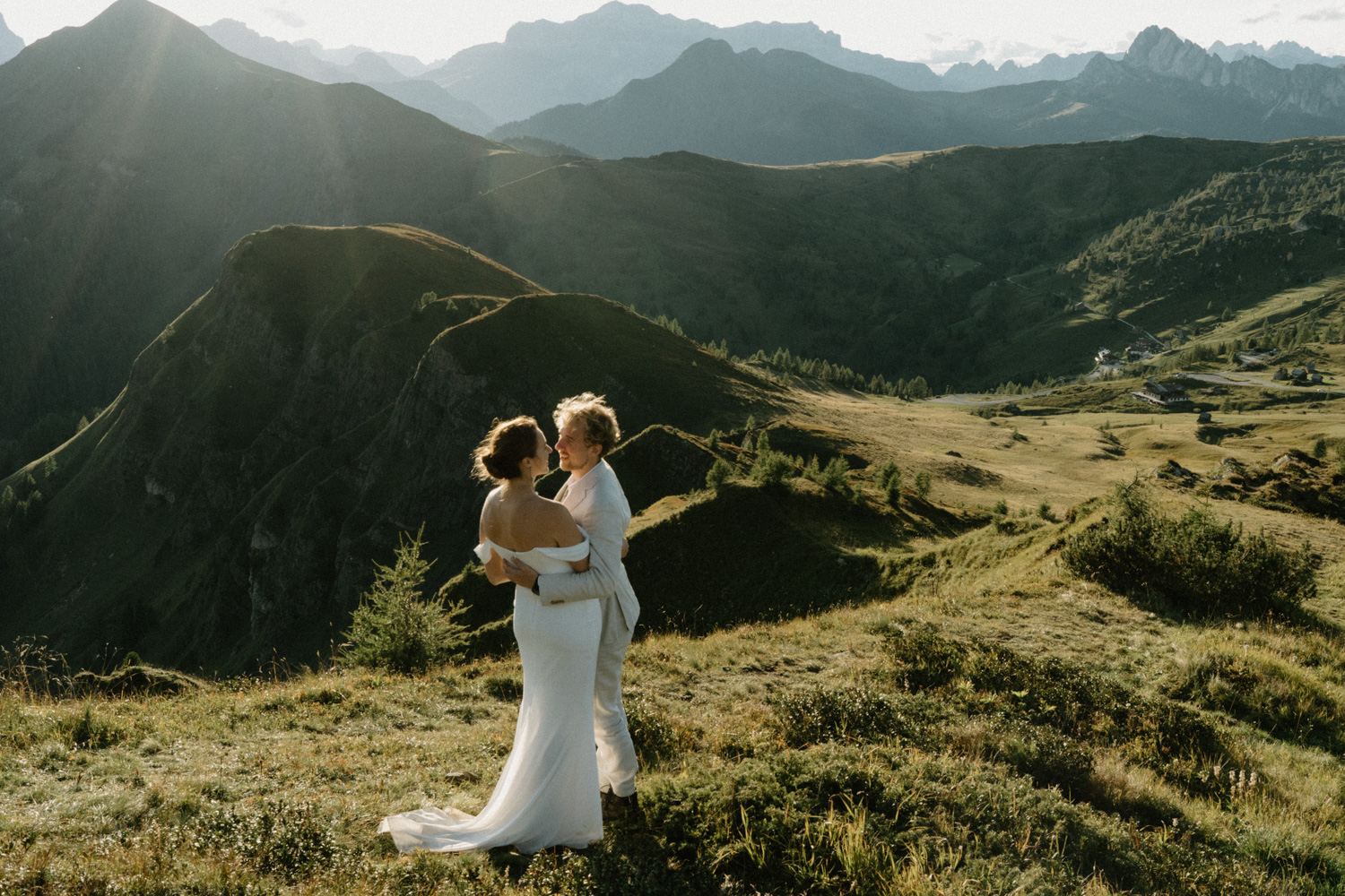 A couple in wedding attire embrace in a green field with the mountains and sunset behind them during their Italian Dolomites elopement. 