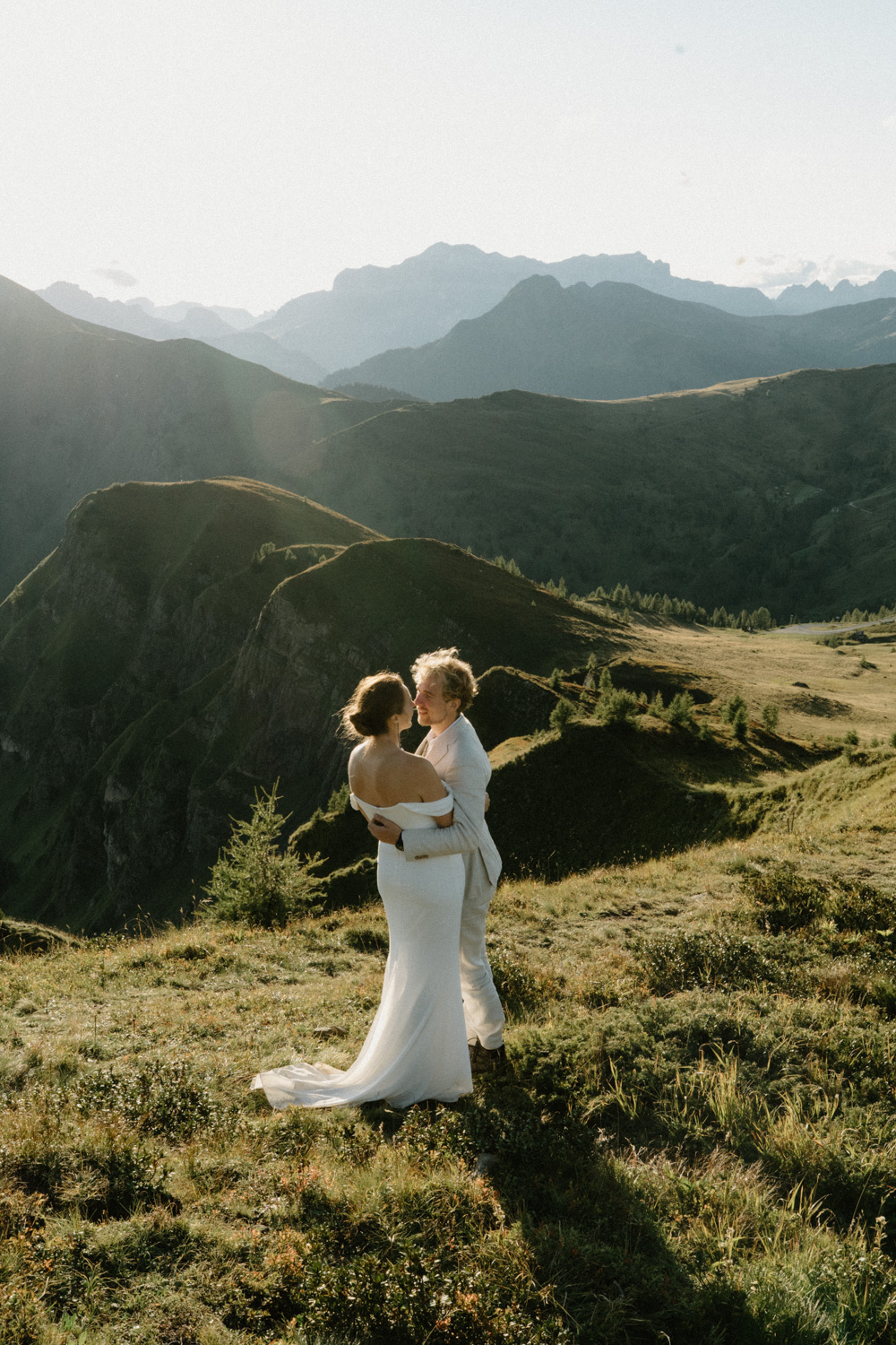 A couple in wedding attire embrace in a green field with the mountains and sunset behind them during their Italian Dolomites elopement. 
