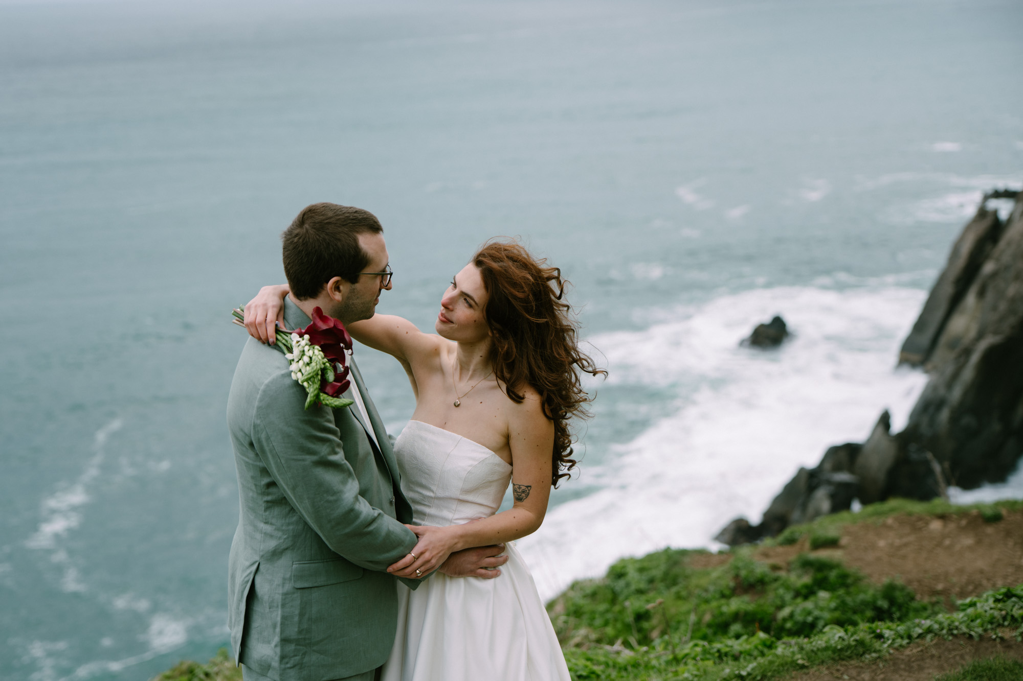 Partners embracing on a dramatic sea cliff at Elk Flats on the Oregon Coast, white dress catching the wind, deep blue Pacific Ocean in the background