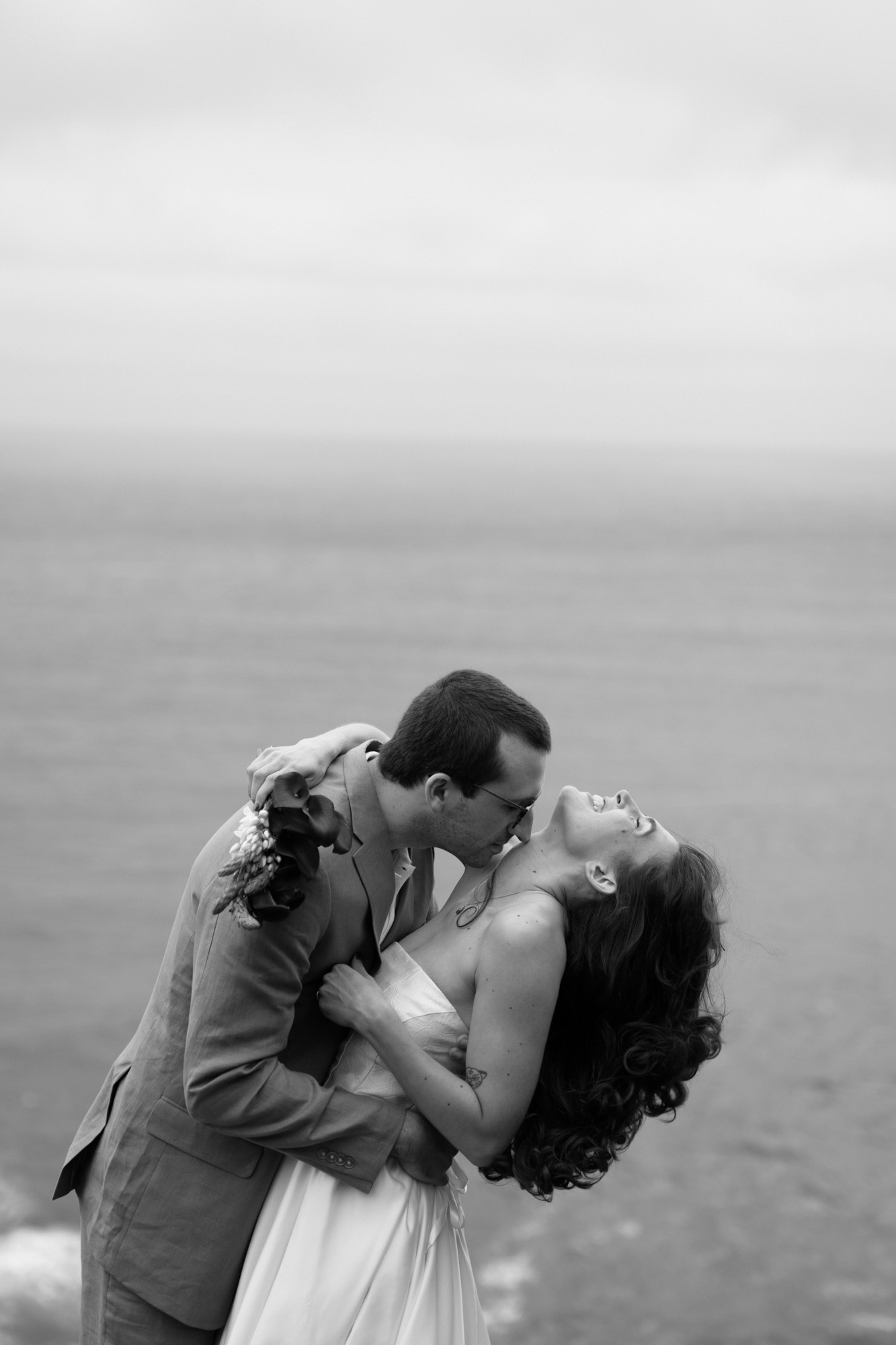 lack and white close-up of a couple kissing on a coastal cliff, laughing mid-kiss with the ocean horizon behind them