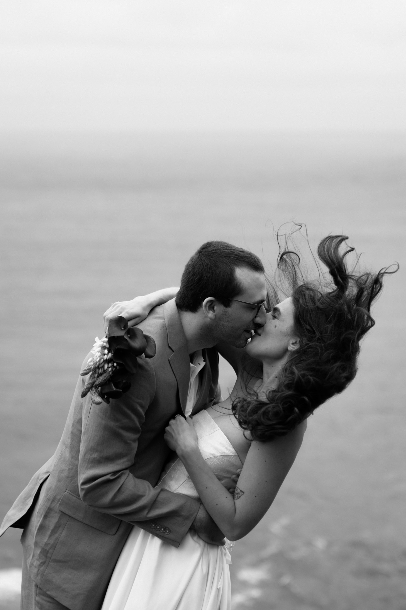 Black and white photo of a couple sharing a first kiss on a windy Oregon Coast cliff, veil and hair flying