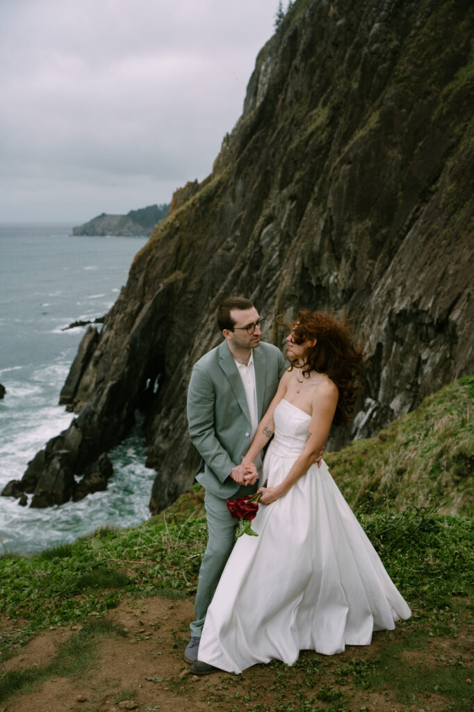 Wide shot of a couple standing at the edge of a sea cliff at Elk Flats, small figures against towering rock formations and the Pacific Ocean