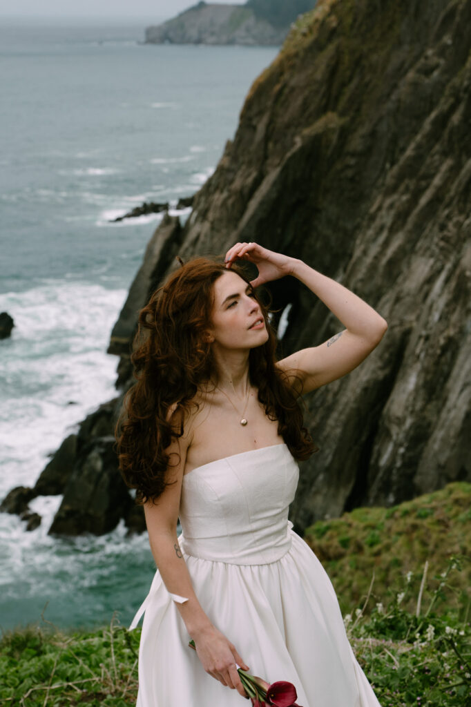 Person in a white dress standing alone on a sea cliff, hand raised to shield from wind, dramatic Oregon Coast cliffs behind them