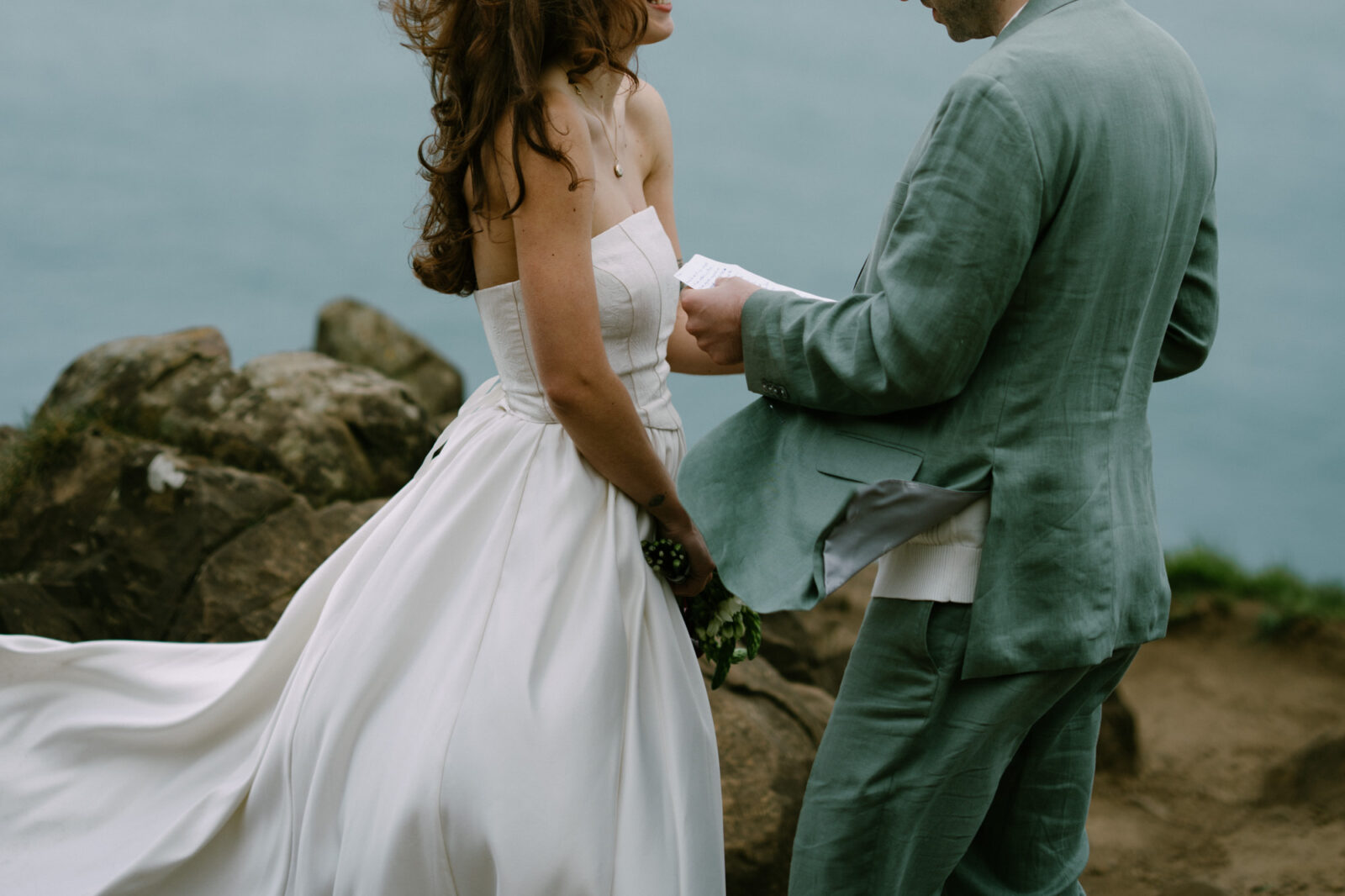 Couple facing each other reading vows at sunset on the Oregon Coast, dress billowing in the wind, rocky sea cliffs behind them