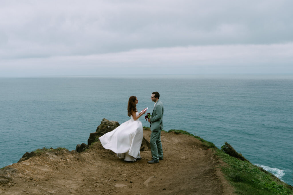 Couple standing on a narrow coastal promontory at Elk Flats reading handwritten vows to each other, vast Pacific Ocean stretching behind them