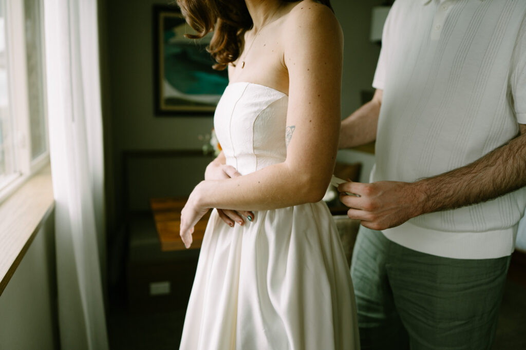 Partner helping fasten the back of a white corset wedding dress at the Stephanie Inn in a room with warm window light