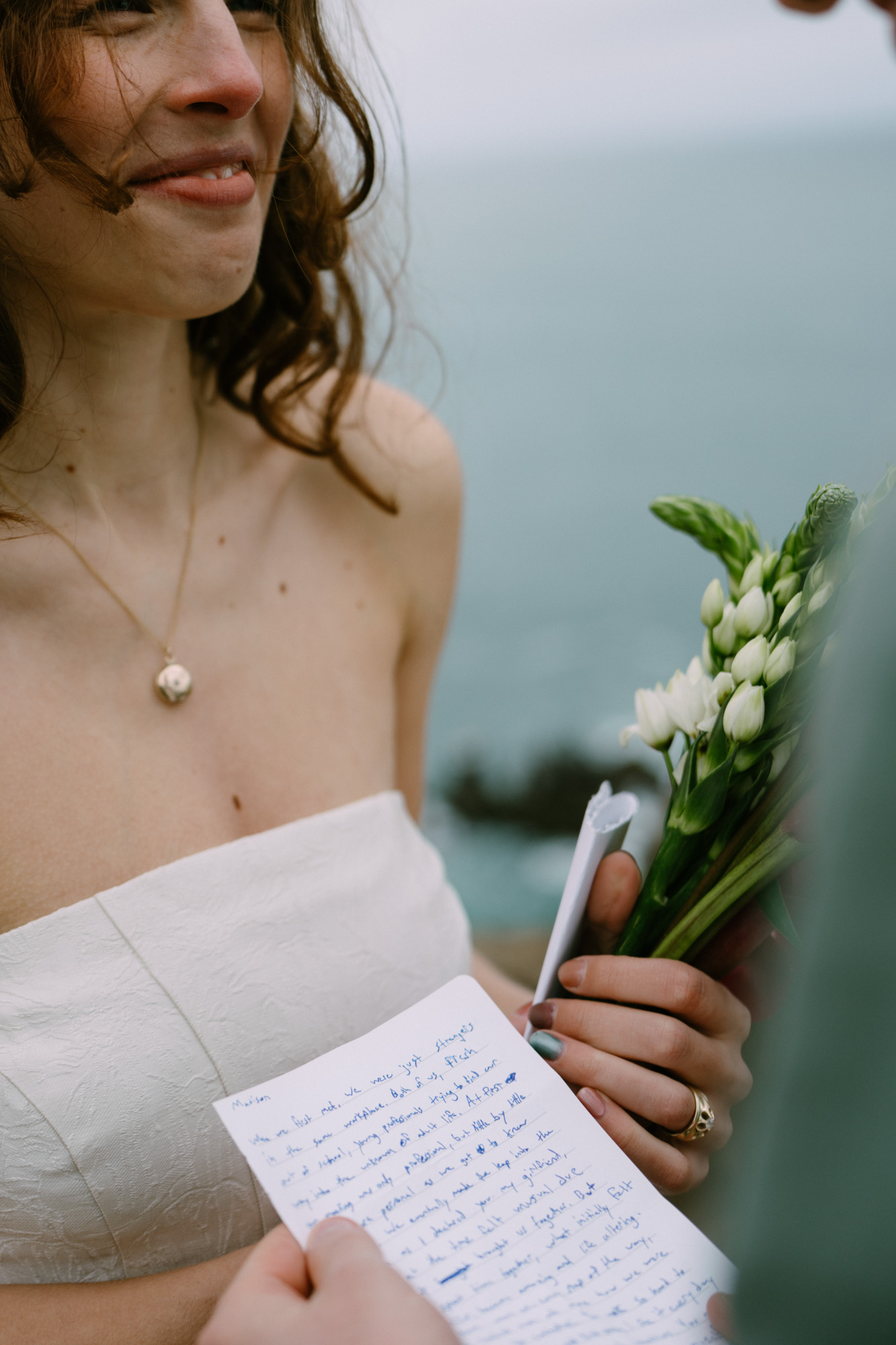 Close-up of handwritten vows on paper, held by hands with a simple ring, coastal light