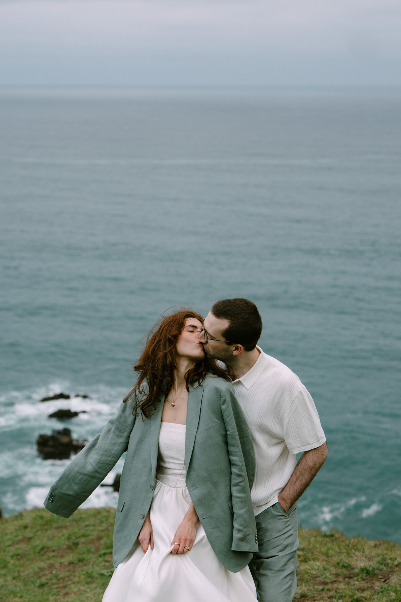 Couple sharing a kiss on a rocky Oregon Coast overlook, hair blowing in the coastal wind
