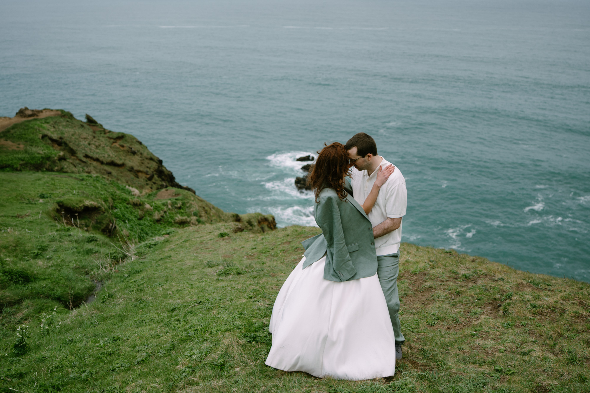 Couple in wedding attire embracing on a grassy cliff at Elk Flats, one partner in a teal jacket, ocean panorama behind them