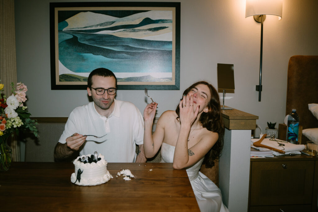 Couple cutting into a small white wedding cake at a wooden table, casual clothes, intimate elopement celebration