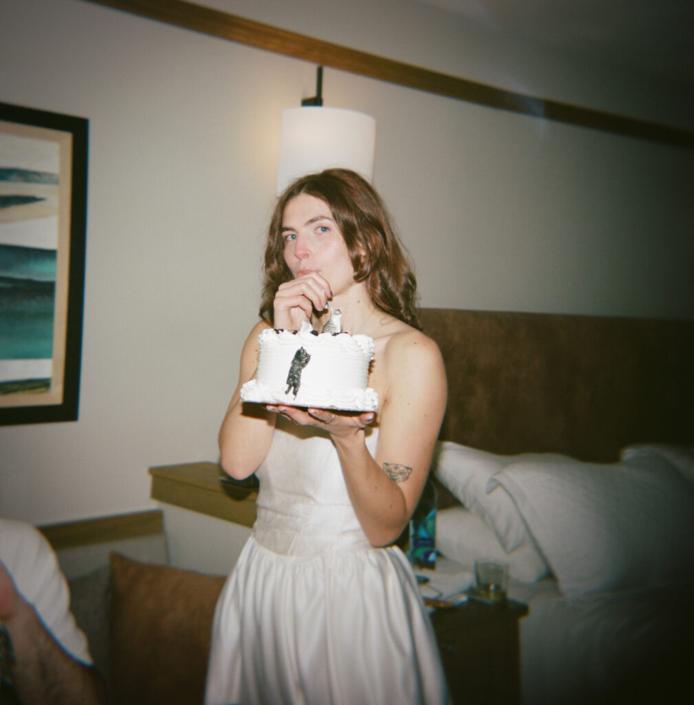 Person in a white wedding dress holding a small wedding cake and eating a bite with a fork, laughing, film photo aesthetic