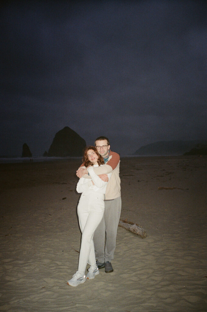 Couple embracing on Cannon Beach at night, Haystack Rock silhouetted in the dark blue sky behind them, film photo