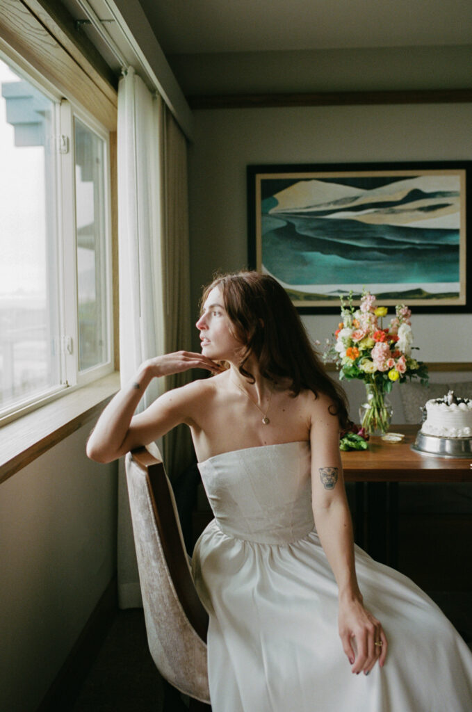 Person in a white wedding dress sitting in a chair by an inn window, looking out with soft contemplative expression, florals and a small cake on the table