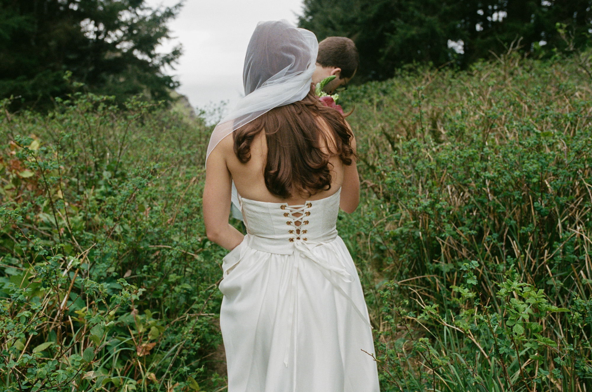 Person in a lace-back wedding dress standing in a wild coastal meadow at Elk Flats, partner leaning in for a kiss, veil lifted by ocean wind