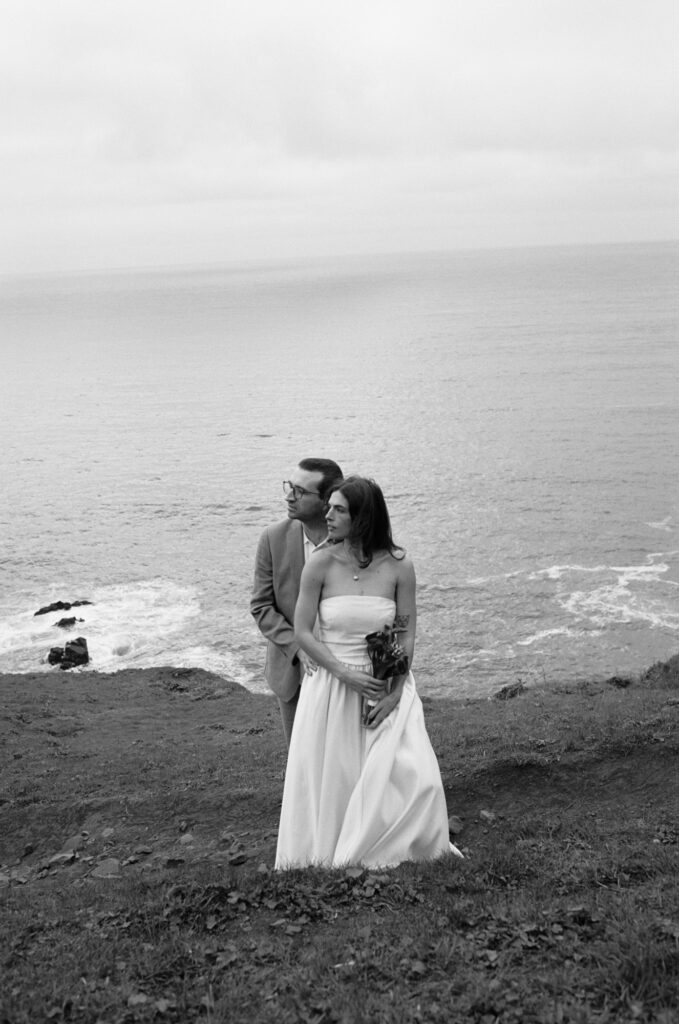 lack and white photo of a couple standing together on a rocky Oregon Coast beach, dressed in wedding attire, ocean behind them