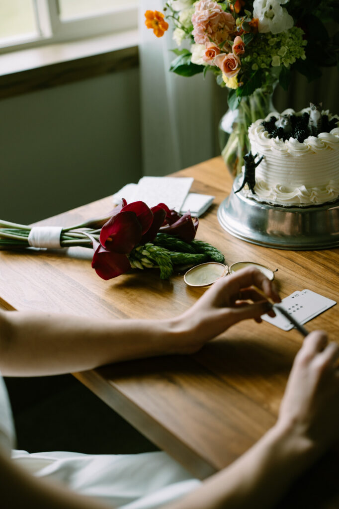lat lay of wedding rings, a small cake, and a loose bouquet of garden flowers including orange ranunculus on a wooden table
