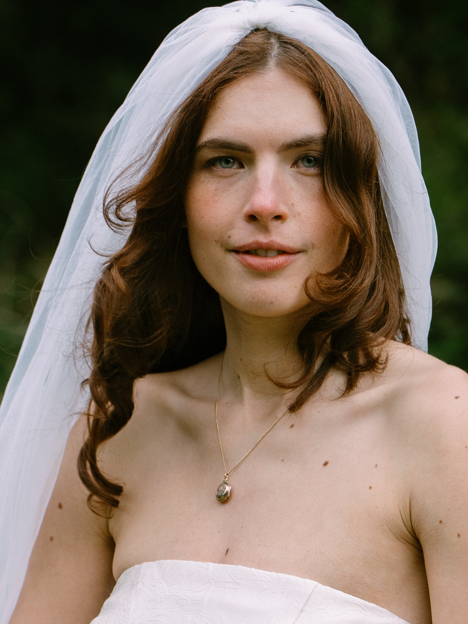 Close-up portrait of a person with auburn hair wearing a soft tulle veil, freckles visible, soft natural light in a coastal forest setting