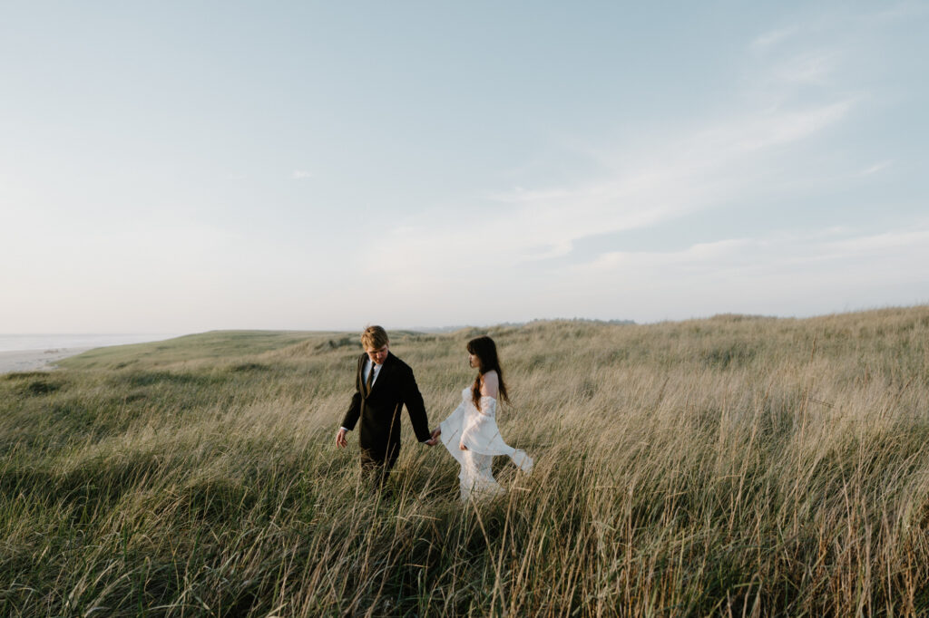 A couple walks hand in hand through tall seagrass during an Oregon coast elopement. 