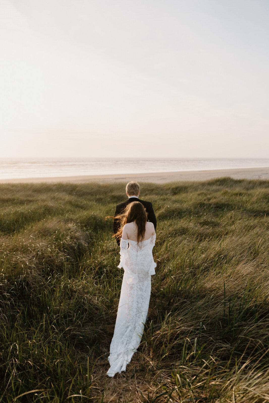A couple walks hand in hand through tall seagrass during an Oregon coast elopement. 
