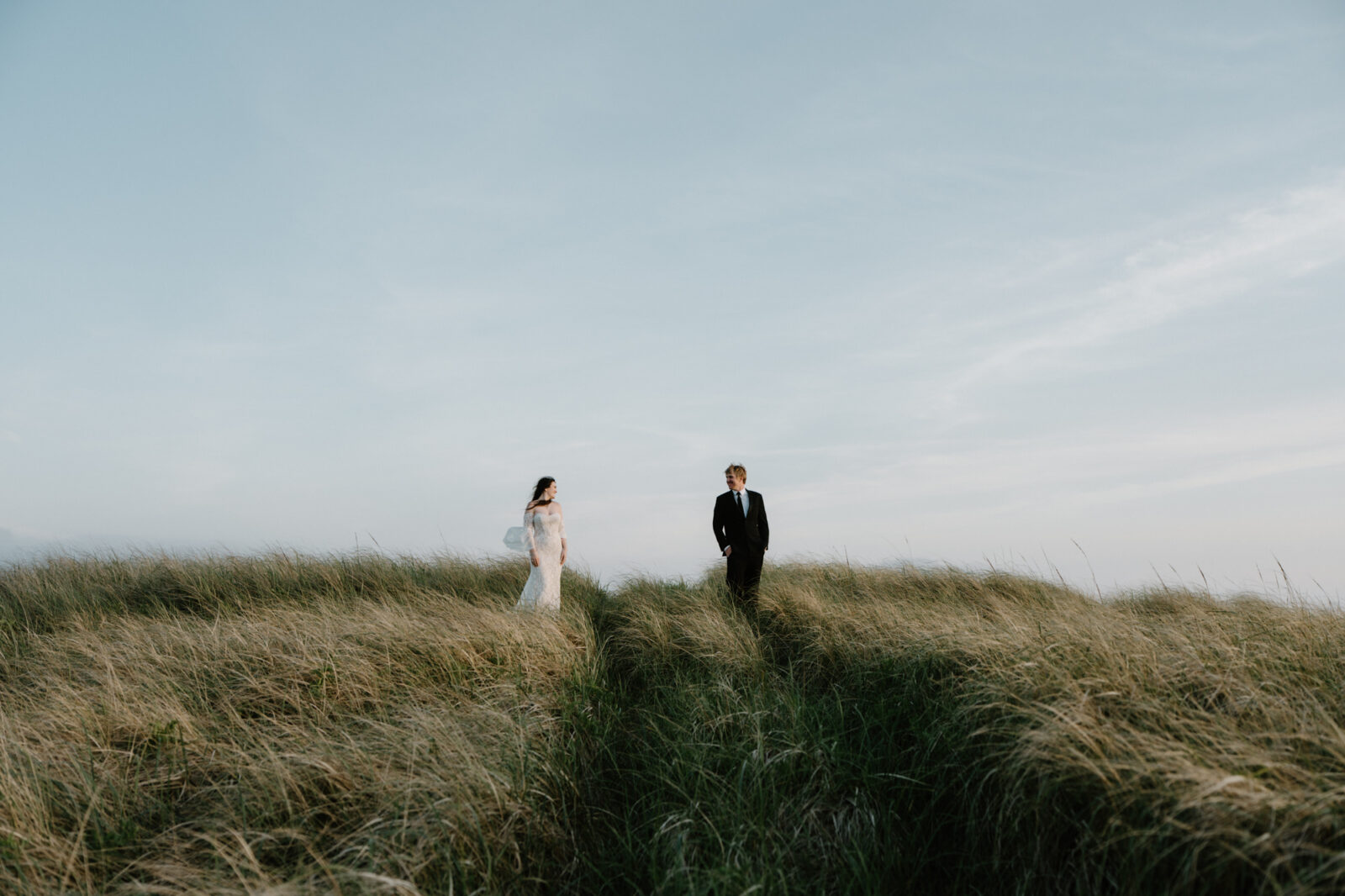 A couple stands on a grassy hill and looks at each other during an Oregon coast elopement. 