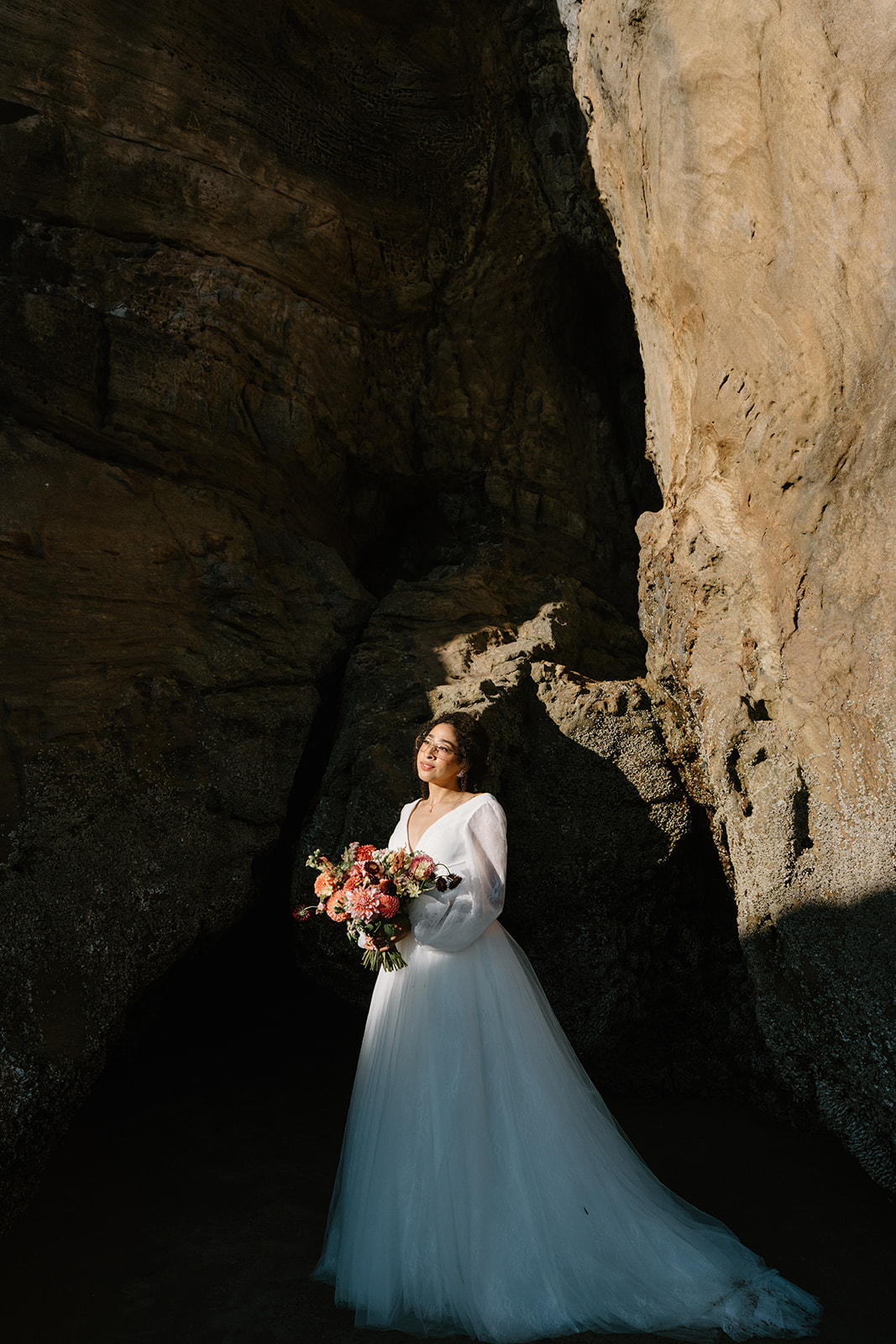 A bride stands next to a red cliff during a Hug Point elopement. 