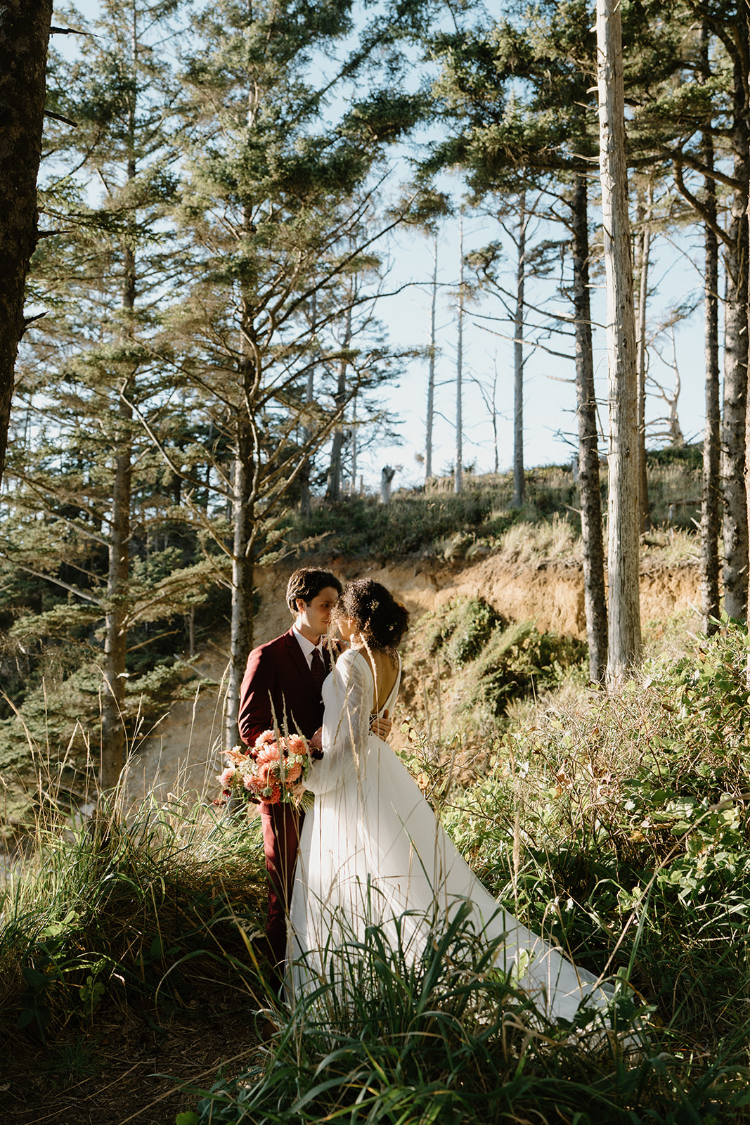 A couple stands on a forested cliff and snuggles close during a Hug Point elopement. 