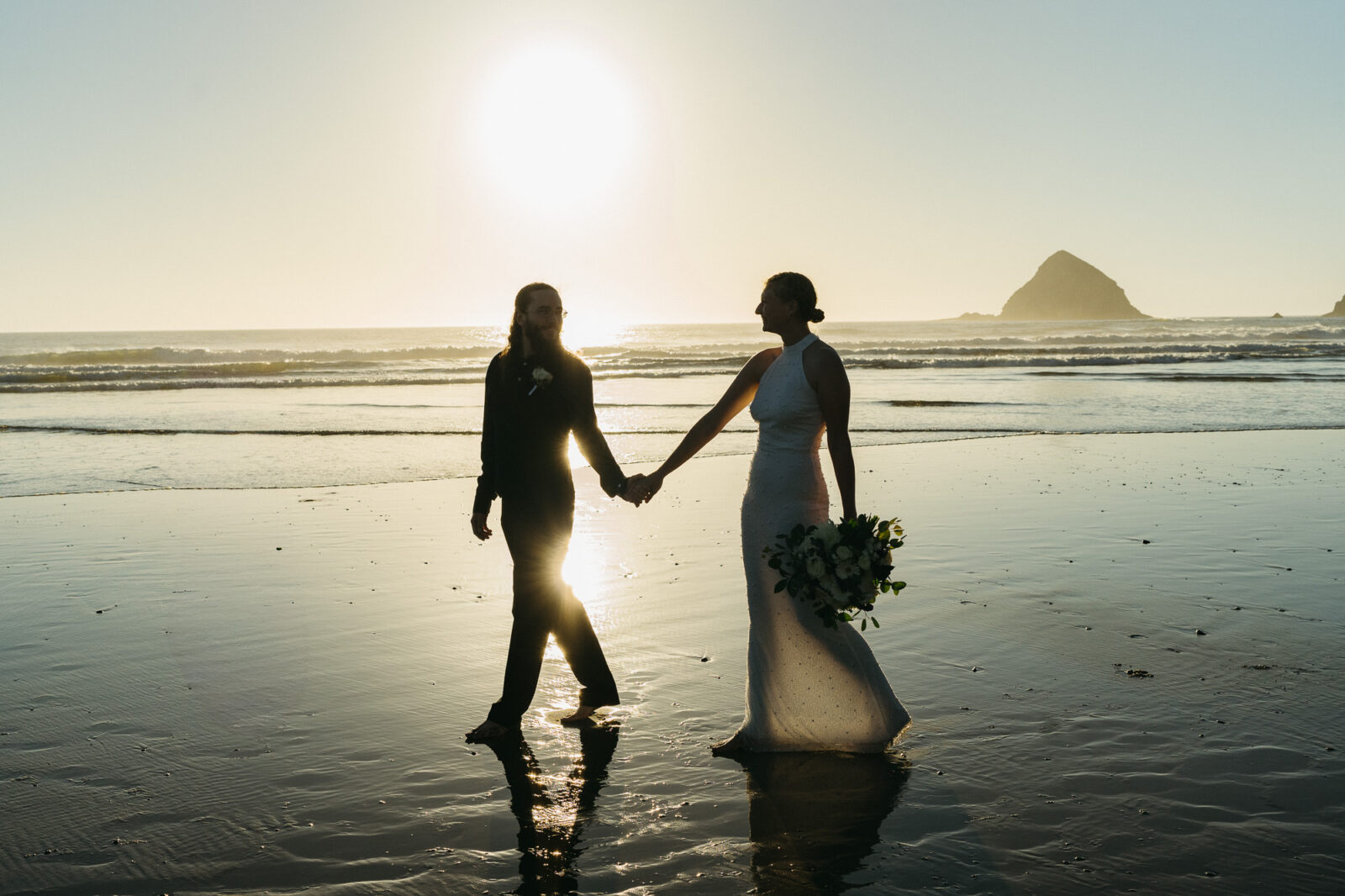 A couple elopes at sunset on the Oregon Coast. There is the ocean, big rocks, and a sandy beach. 