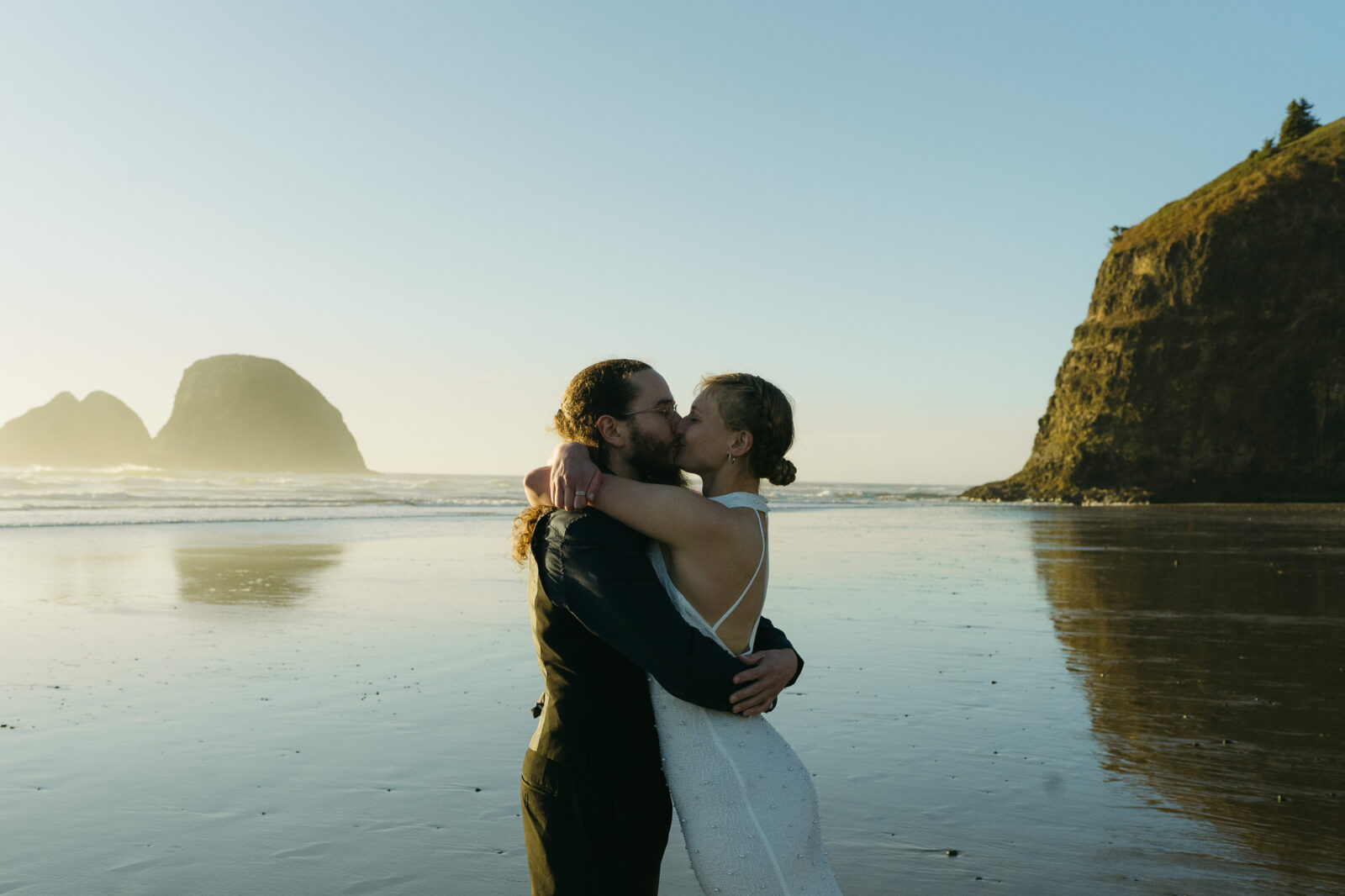 A couple elopes at sunset on the Oregon Coast. There is the ocean, big rocks, and a sandy beach. 