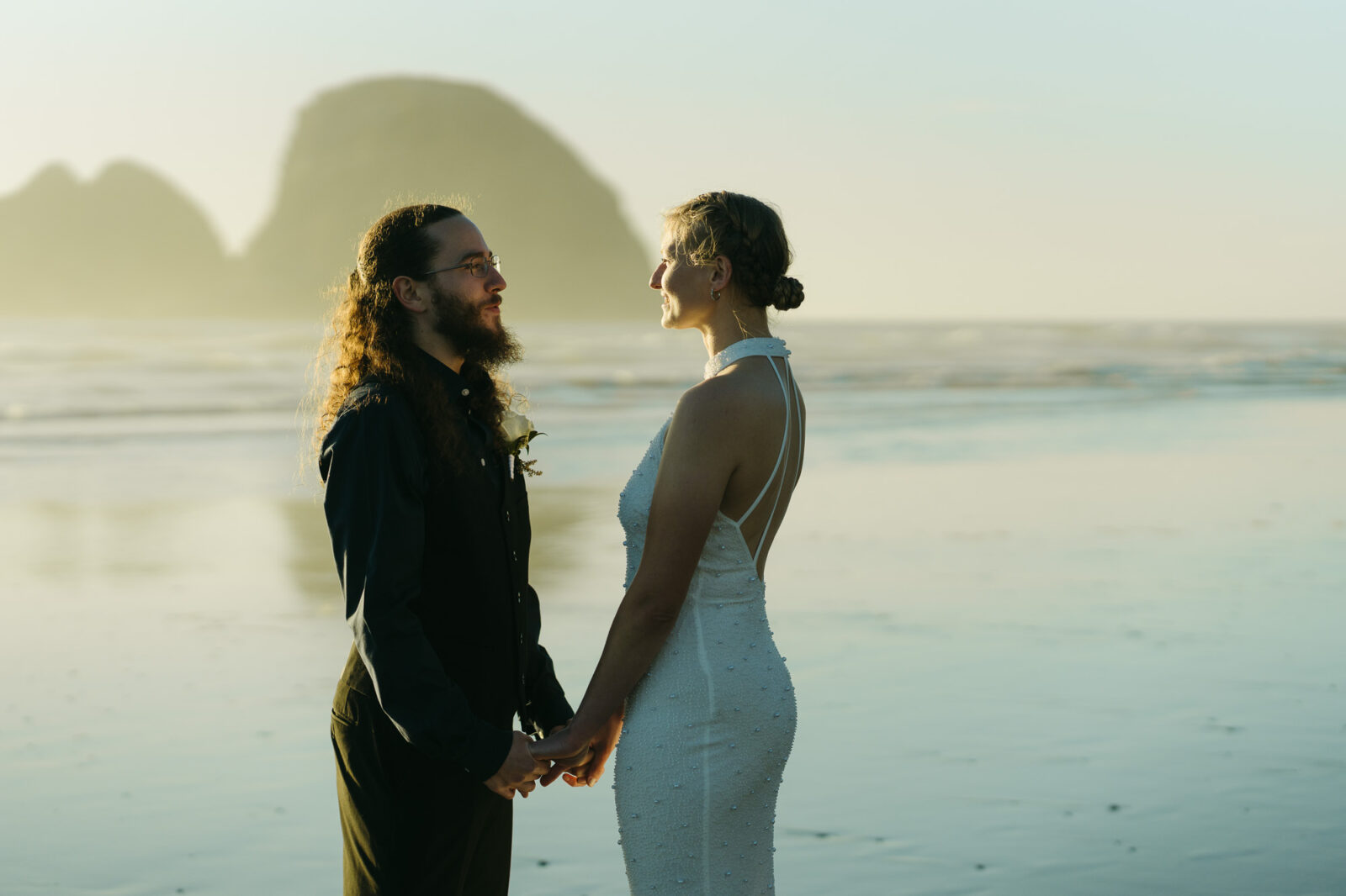 A couple elopes at sunset on the Oregon Coast. There is the ocean, big rocks, and a sandy beach. 