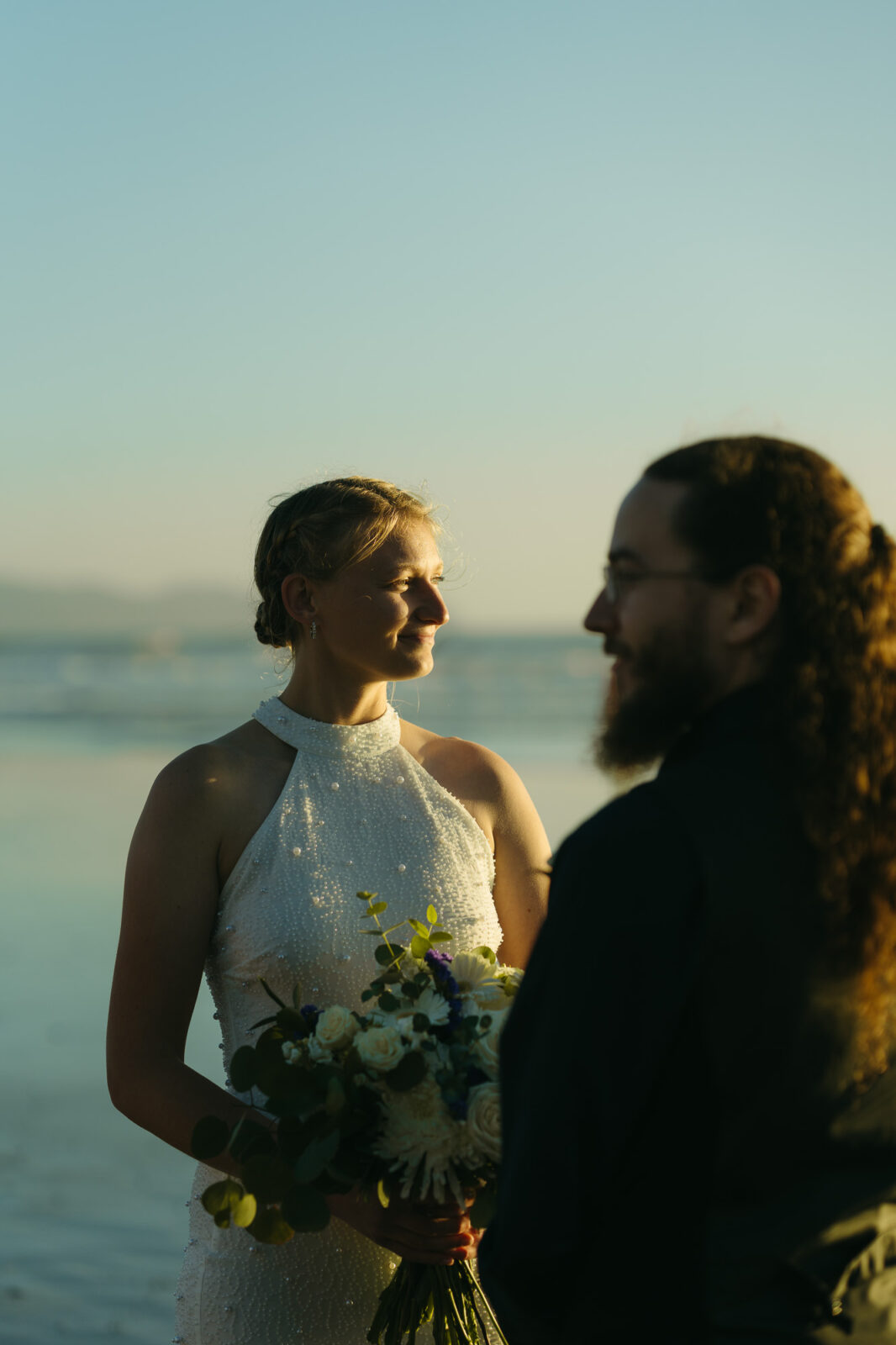 A couple elopes at sunset on the Oregon Coast. There is the ocean, big rocks, and a sandy beach. 