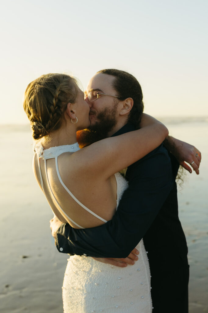 A couple elopes at sunset on the Oregon Coast. There is the ocean, big rocks, and a sandy beach. 