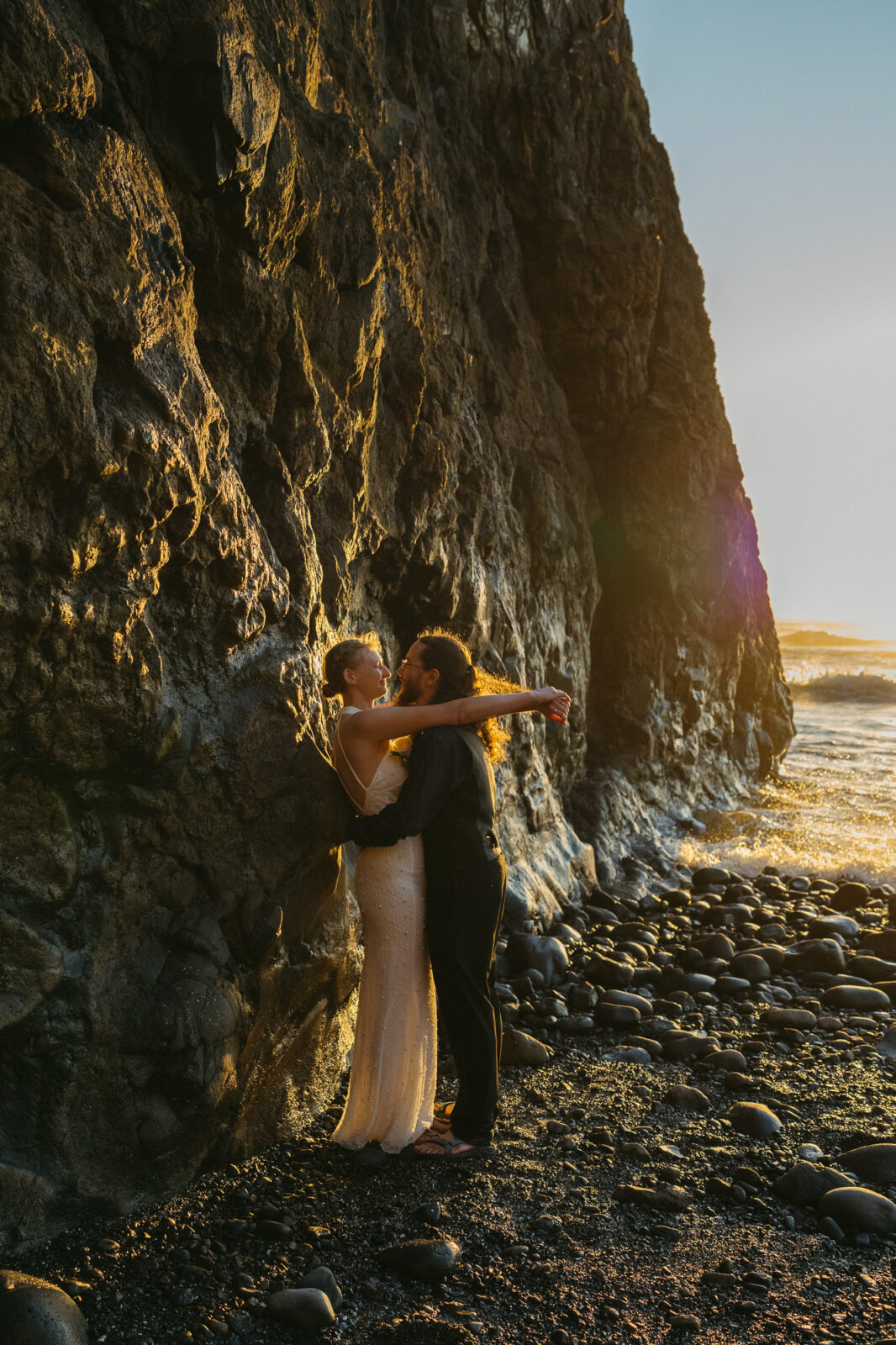 A couple elopes at sunset on the Oregon Coast. There is the ocean, big rocks, and a sandy beach. 