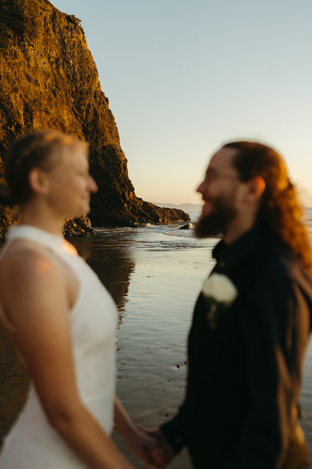 A couple elopes at sunset on the Oregon Coast. There is the ocean, big rocks, and a sandy beach. 