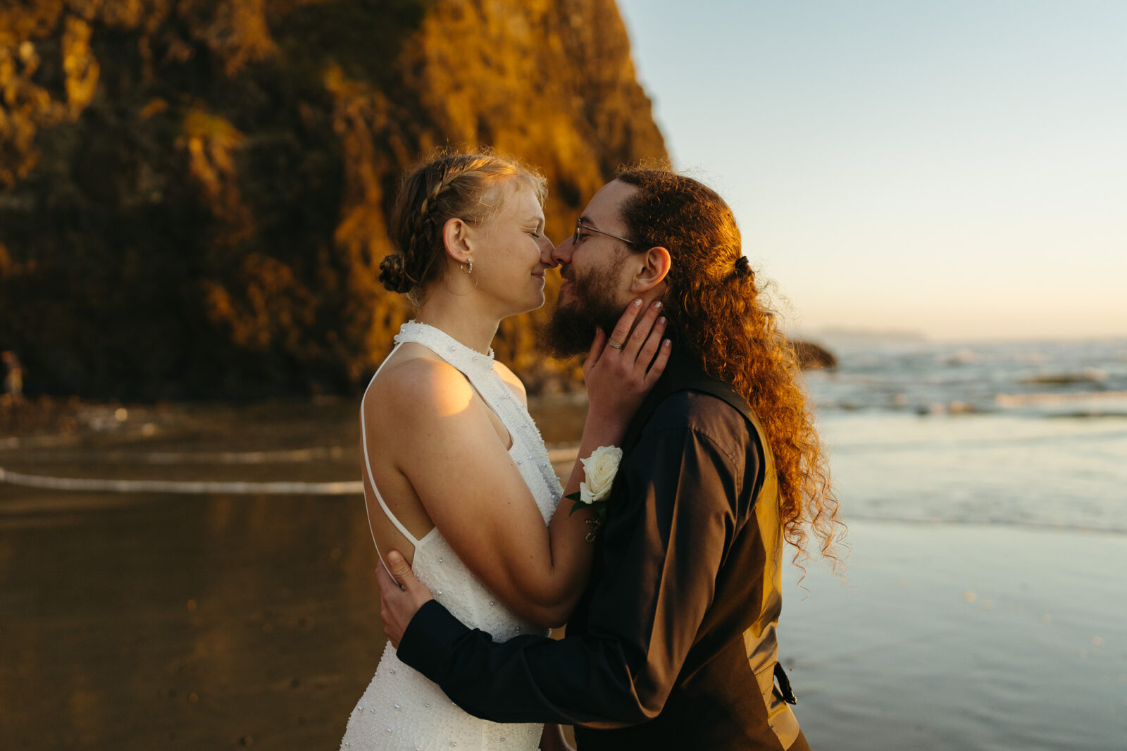 A couple elopes at sunset on the Oregon Coast. There is the ocean, big rocks, and a sandy beach. 