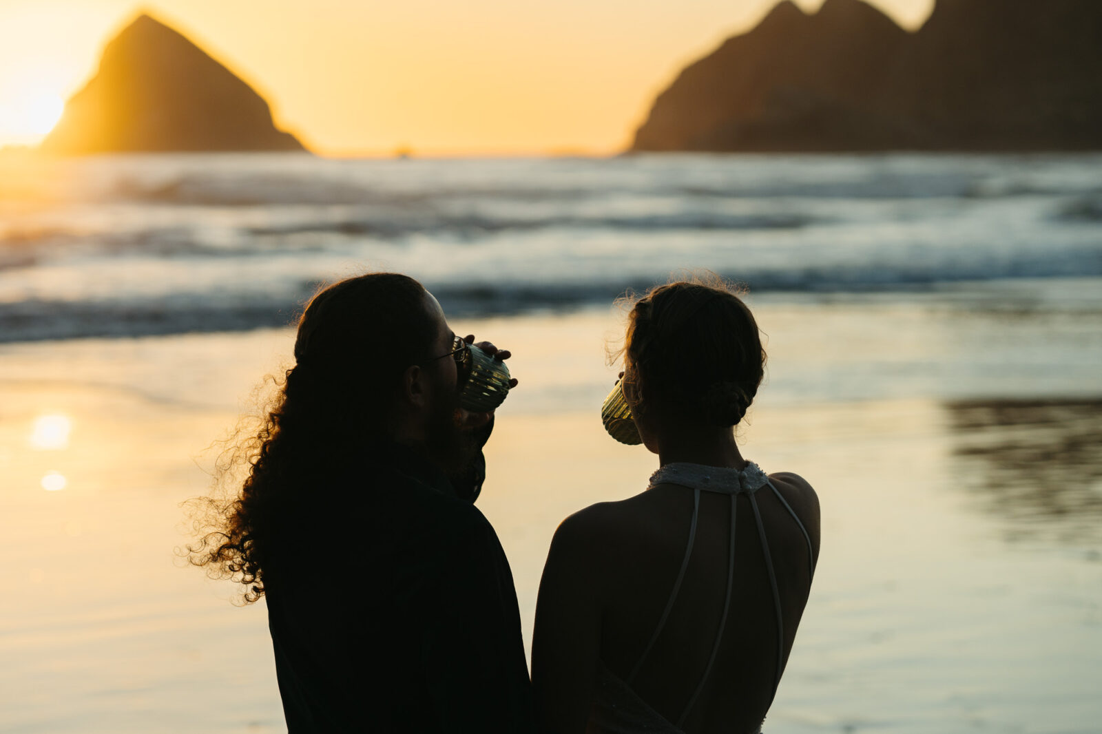 A couple elopes at sunset on the Oregon Coast. There is the ocean, big rocks, and a sandy beach. 