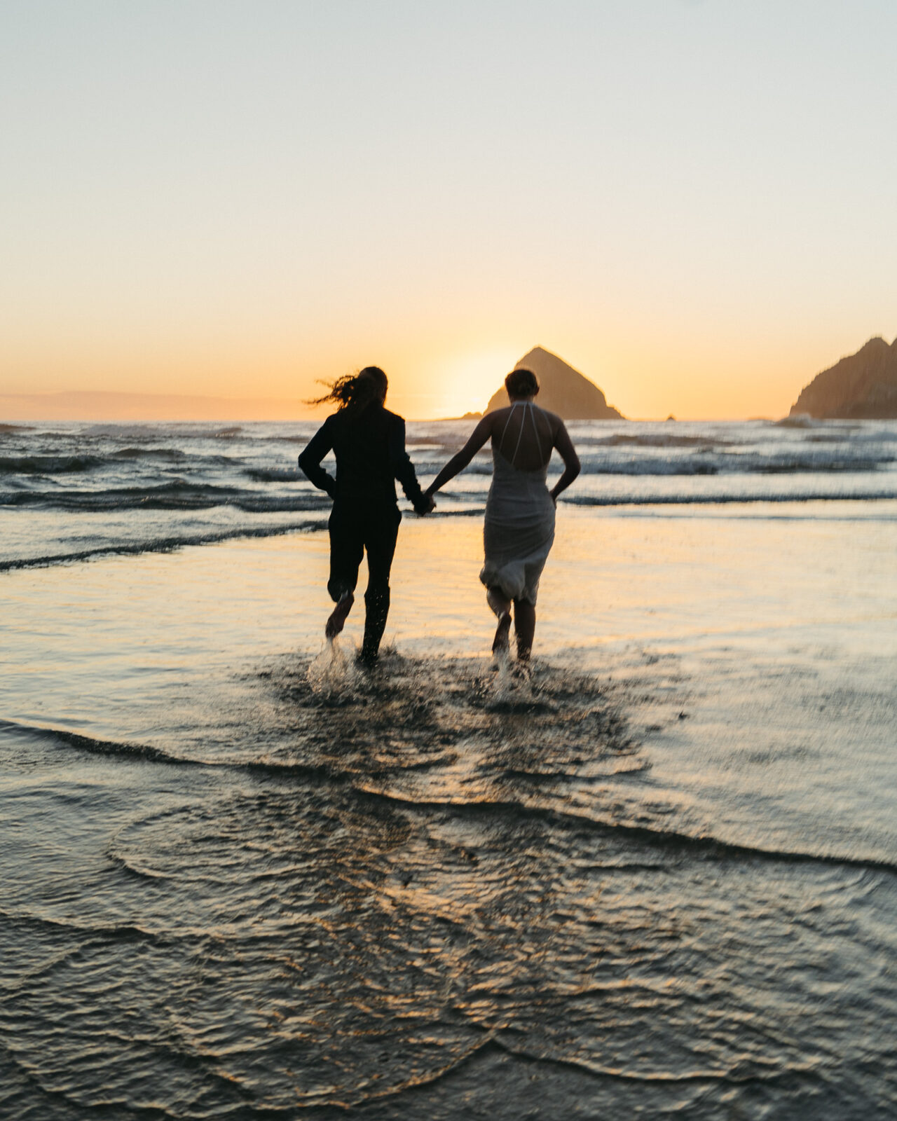 A couple elopes at sunset on the Oregon Coast. There is the ocean, big rocks, and a sandy beach. 