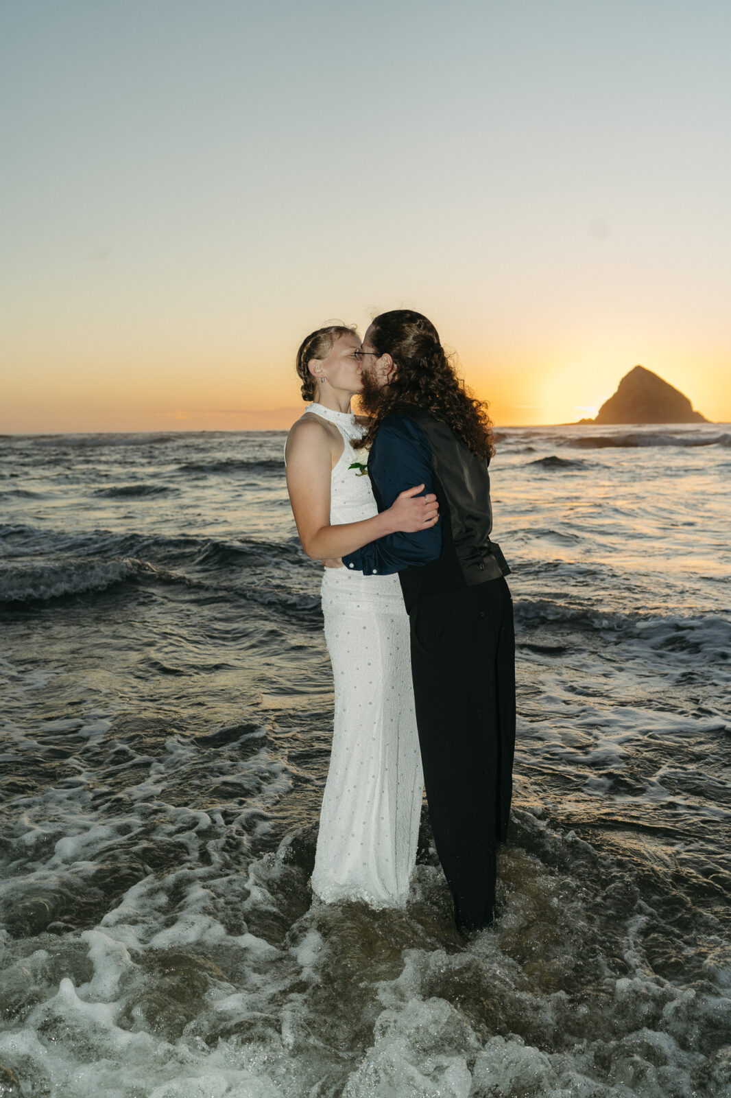 A couple elopes at sunset on the Oregon Coast. There is the ocean, big rocks, and a sandy beach. 