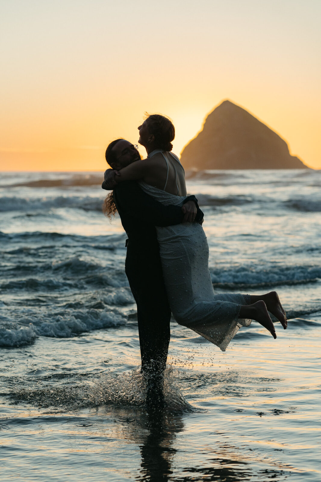 A couple elopes at sunset on the Oregon Coast. There is the ocean, big rocks, and a sandy beach. 